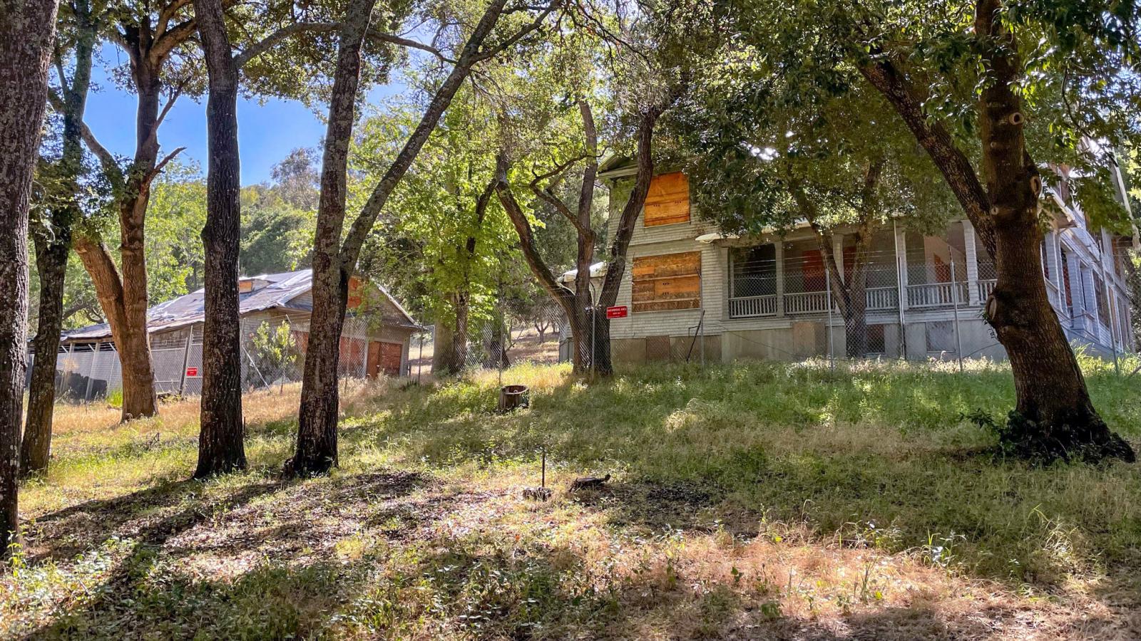 Historic house with boarded-up windows and outbuilding visible through oak trees
