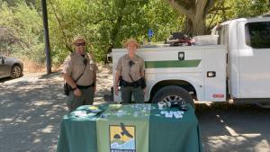 Two Midpen Rangers stand behind a table with Midpen stickers, pins, and other branded items that they are giving away to members of the public